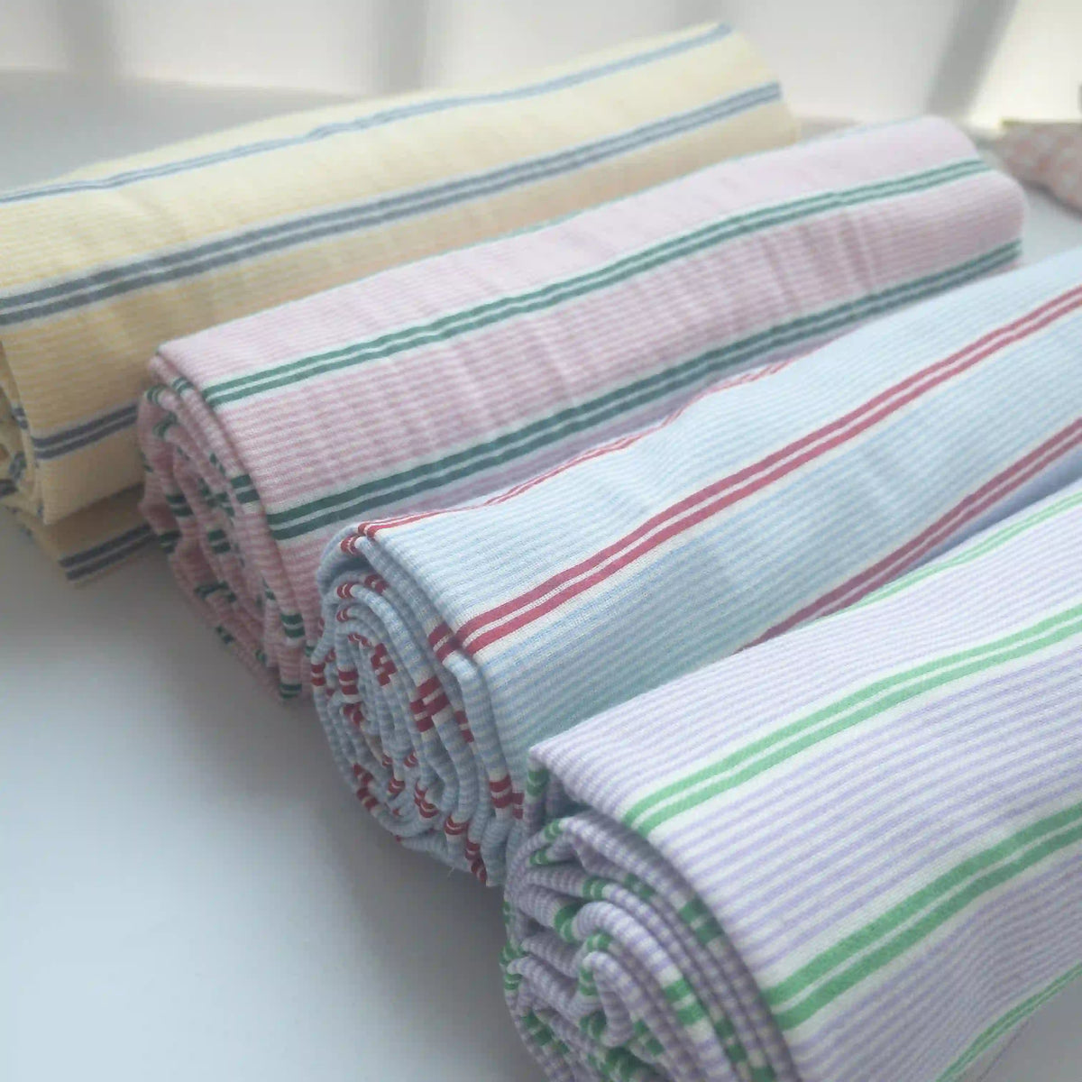 Four rolls of Longan Craft's Striped Printed Gauze Fabric by the yard—breathable, double 100% cotton with pastel yellow, pink, blue, green, and red stripes—are neatly displayed on a white surface in natural light.
