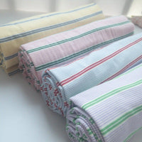 Four rolls of Longan Craft's Striped Printed Gauze Fabric by the yard—breathable, double 100% cotton with pastel yellow, pink, blue, green, and red stripes—are neatly displayed on a white surface in natural light.