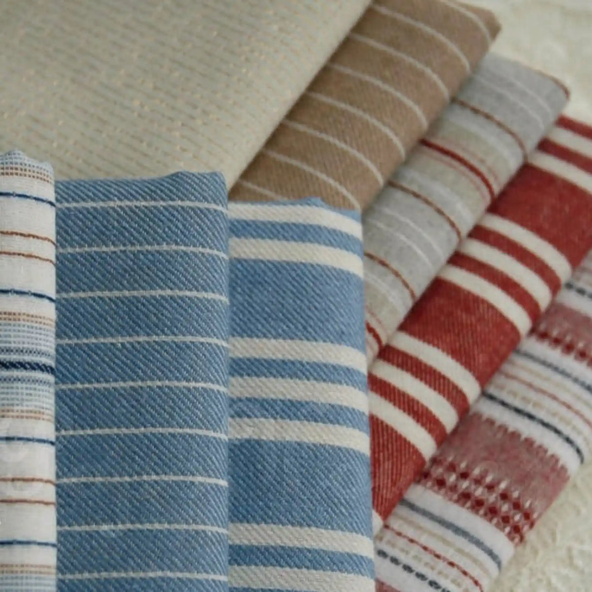 A neatly stacked selection of Longan Craft’s Striped Printed Linen Fabric By The Yard showcases a variety of colorful stripe patterns and textures in beige, blue, and red. Each cotton linen blend fabric piece highlights the natural beauty and breathability perfect for dresses.