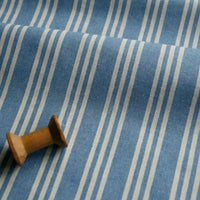 A wooden spool rests on Longan Craft’s Striped Printed Linen Fabric by the Yard, a breathable cotton-linen blend featuring a colorful stripe pattern with evenly spaced vertical white stripes. The fabric appears soft and slightly wrinkled.