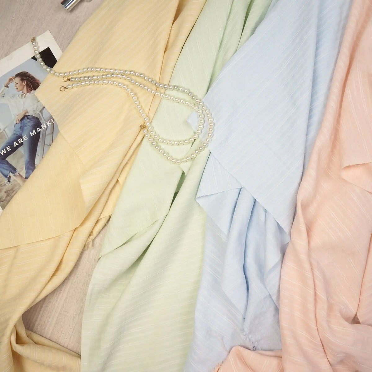 Pastel yellow, green, blue, and peach fabrics are laid out with a pearl necklace and a photo atop the yellow Striped Rayon Dobby Fabric By The Yard from Longan Craft, a polyester jacquard perfect for blouses.