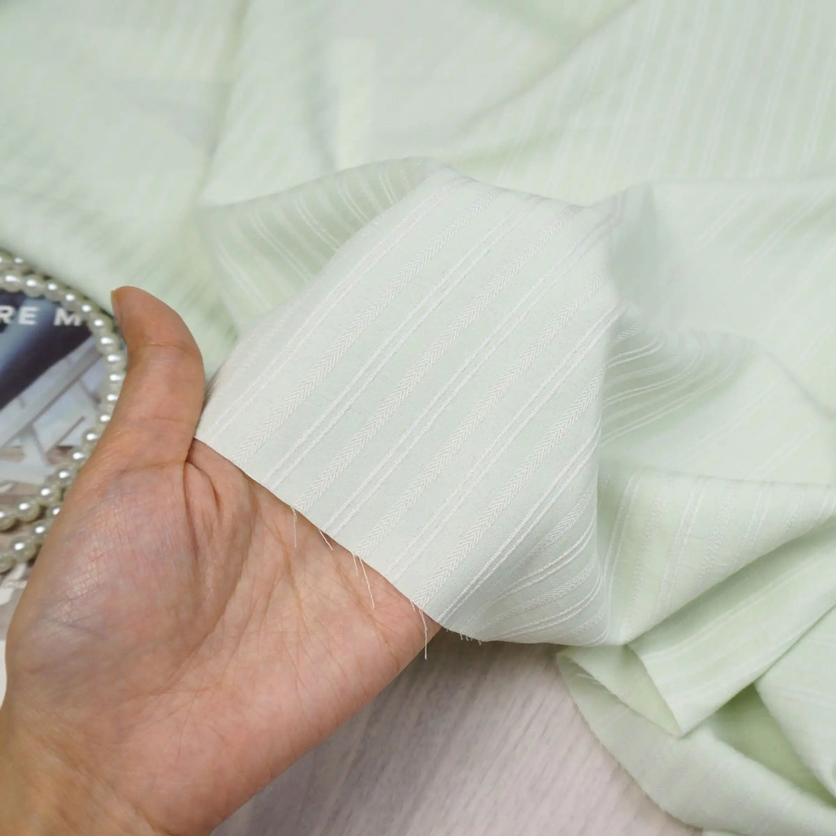 A hand displays Longan Craft's Striped Rayon Dobby Fabric by the yard, highlighting its dobby jacquard texture. Polyester material ideal for blouses. Pearls and a book or magazine appear in the background.