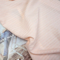 A soft, light pink Striped Rayon Dobby Fabric by Longan Craft, featuring thin white stripes, is draped over a magazine showing someone in blue jeans and a beige sweater seated on a chair. Ideal for blouses.