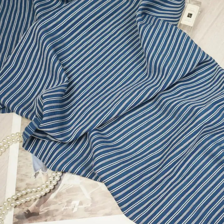 A blue Striped Rayon Dobby Fabric by the yard from Longan Craft, featuring thin white stripes, is elegantly displayed with pearls and a clear perfume bottle on a light surface—ideal jacquard textile for blouses and crafts.