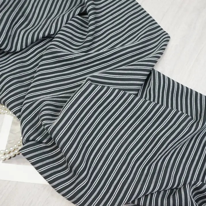 A close-up of Longan Craft Striped Rayon Dobby Fabric By The Yard shows thin white vertical stripes on black polyester jacquard, slightly wrinkled and draped over a light surface, with a strand of pearls partly visible beneath the fabric.