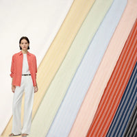 A woman in a red and white striped shirt with white trousers stands before pastel, diagonal swatches of Longan Craft Striped Rayon Dobby Fabric by the yard—an ideal polyester jacquard dobby textile for blouse-making inspiration.