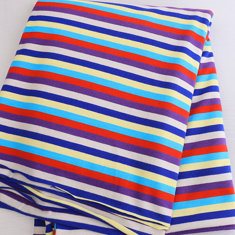 A folded Longan Craft Striped Jersey Fabric By The Yard in cotton spandex, featuring purple, red, yellow, and light blue diagonal stripes on white, rests on a light surface—perfect for DIY short sleeves.