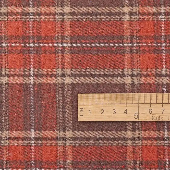 A wooden ruler displaying centimeter measurements from 1 to 7 rests atop Longan Craft’s Thick Plaid Mammoth Flannel Fabric By The Yard. This heavyweight cotton brushed flannel features a classic red and brown plaid pattern with intersecting beige, white, and orange stripes—ideal for anyone seeking warm, cozy apparel fabric sold by the yard.