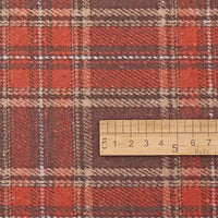 A wooden ruler displaying centimeter measurements from 1 to 7 rests atop Longan Craft’s Thick Plaid Mammoth Flannel Fabric By The Yard. This heavyweight cotton brushed flannel features a classic red and brown plaid pattern with intersecting beige, white, and orange stripes—ideal for anyone seeking warm, cozy apparel fabric sold by the yard.