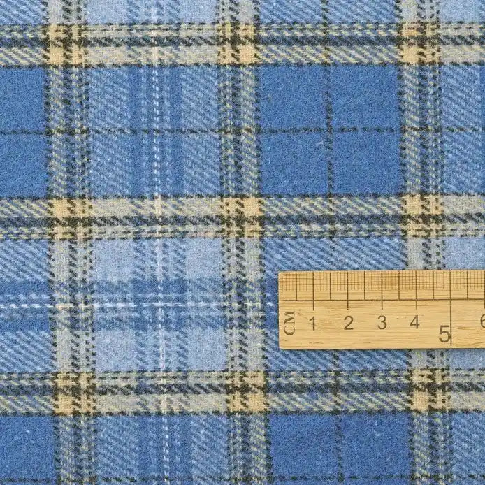A wooden ruler rests atop Longan Craft’s Thick Plaid Mammoth Flannel Fabric By The Yard, a heavyweight cotton brushed flannel featuring light and dark blue, white, and beige checkered stripes—ideal for accurate measurements or selecting this warm apparel fabric by the yard.