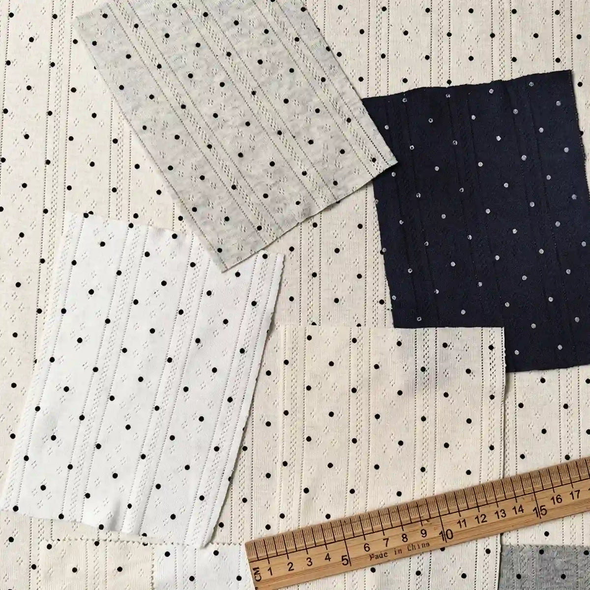 Four swatches of Tiny Polka Dot Print Pointelle Fabric By The Yard in white, cream, and navy polka dot patterns arranged near a wooden ruler.