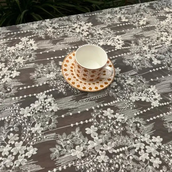 A white teacup with a patterned saucer rests on Longan Craft's Vintage Floral Embroidery Lace Fabric By The Yard, showcasing delicate white botanical designs on gray—perfect cotton lace material for dress creations.