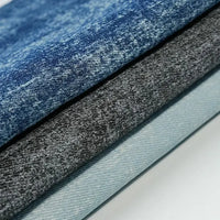 A stack of Washed Denim Fabric By The Yard from Longan Craft, featuring premium soft vintage cotton denim in blue, dark gray, and light blue tones, showcases the stylish and durable appeal of this quality textile for jeans.