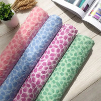 Four rolls of patterned fabric in pink, blue, purple, and green—including the Watercolor Pebble Print Seersucker Fabric by Longan Craft—are displayed on a light wooden surface next to a small potted plant, wheat stalks, and an open magazine.