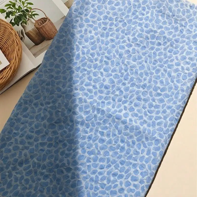 The Watercolor Pebble Print Seersucker Fabric by the yard from Longan Craft, made of 100% cotton and featuring an abstract pebble pattern in light blue tones, is displayed on a beige surface beside an open magazine and a woven basket with a potted plant.