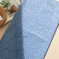 The Watercolor Pebble Print Seersucker Fabric by the yard from Longan Craft, made of 100% cotton and featuring an abstract pebble pattern in light blue tones, is displayed on a beige surface beside an open magazine and a woven basket with a potted plant.