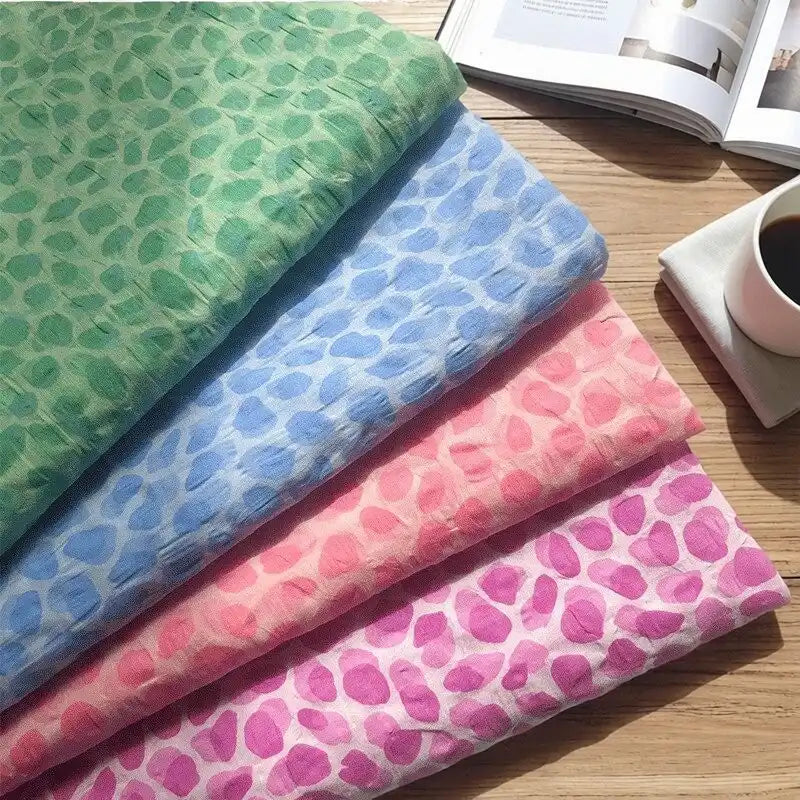 Four folded pieces of Longan Craft Watercolor Pebble Print Seersucker Fabric By The Yard in green, blue, pink, and purple are displayed on a wooden surface beside an open magazine and a cup of coffee on a white saucer.