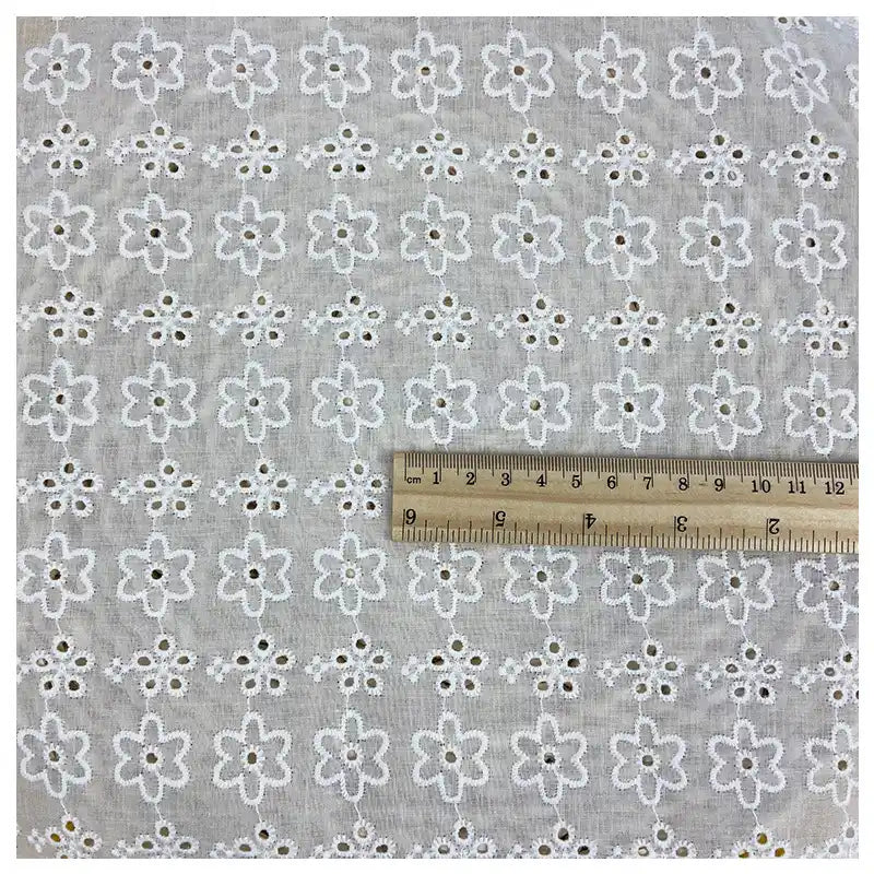 The Longan Craft White Floral Eyelet Embroidered Voile Fabric by the yard is a lightweight, breathable 100% cotton material in light gray with white floral hollow-out embroidery. A wooden ruler in centimeters and inches shows the scale on the fabric.