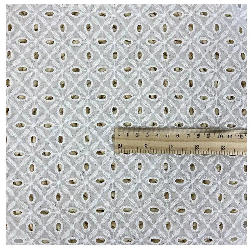 A sample of Longan Craft's White Floral Eyelet Embroidered Voile Fabric, 100% cotton with geometric circular and diamond embroidery, is shown next to a ruler for scale; the piece measures about 8 inches wide. Ideal for dressmaking.