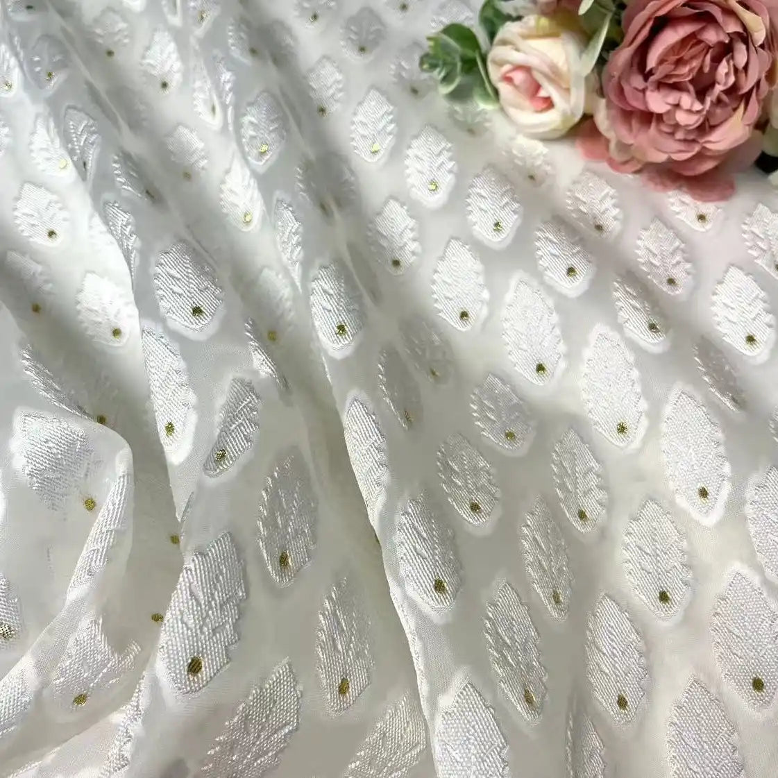 Longan Craft's White Leaf Pattern Organza Fabric by the yard features a subtle botanical jacquard design with gold dots. Artificial pink and cream roses with green leaves rest gently on this sheer organza, ideal for clothing projects.