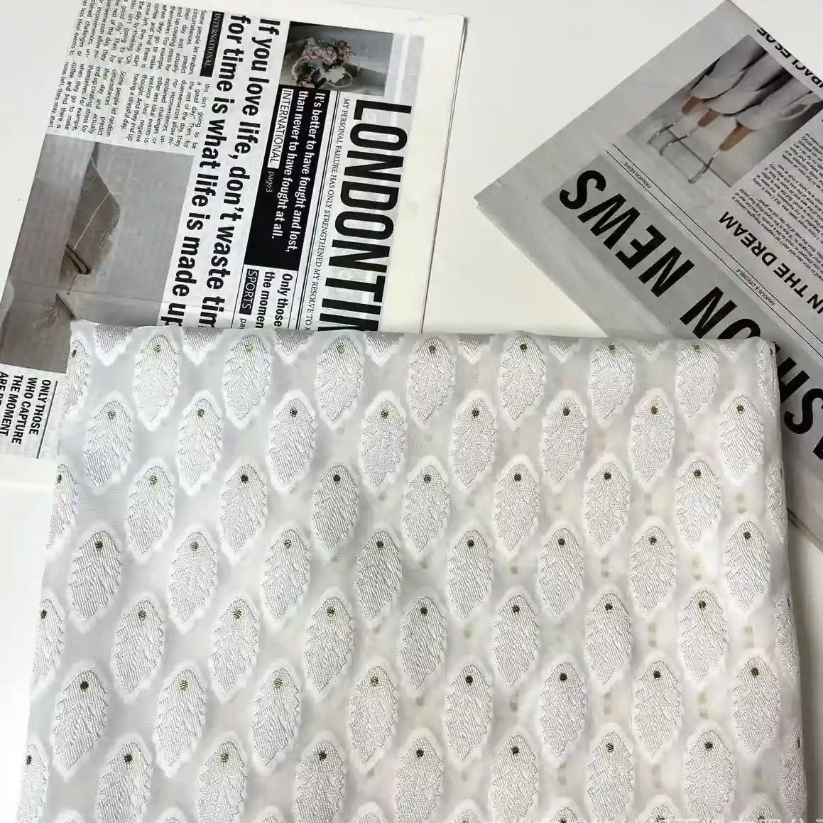 Longan Craft’s White Leaf Pattern Organza Fabric By The Yard, a botanical jacquard material for clothing, is displayed on a white surface with newspaper sheets adorned with bold headlines and fashion images.