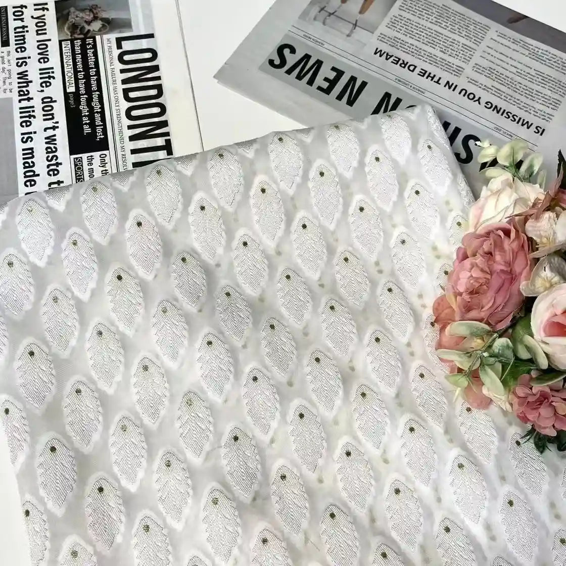 A piece of Longan Craft’s White Leaf Pattern Organza Fabric By The Yard is displayed on a table beside pink and white flowers and newspapers, showcasing its botanical jacquard texture—ideal organza material for clothing.