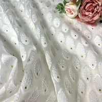 The White Leaf Pattern Organza Fabric by Longan Craft features an embroidered leaf design and gold dots, draped softly. Pink and white artificial flowers rest on the fabric’s upper right corner, highlighting its elegant botanical jacquard pattern.