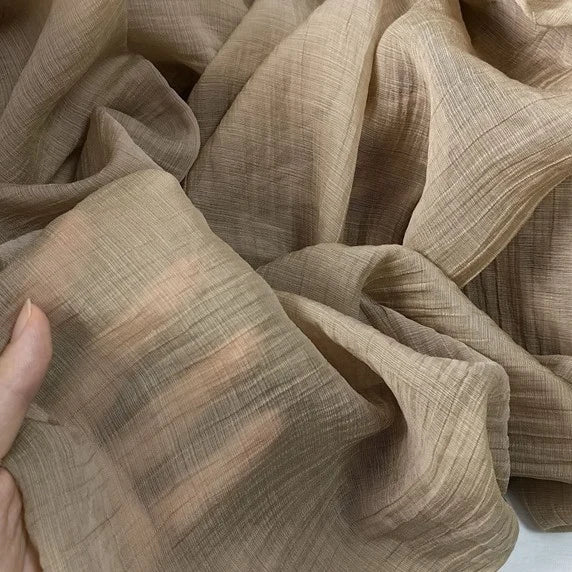 A close-up of a person holding Longan Craft's Wrinkled Lyocell Fabric by the yard, a light brown, sustainable and breathable material with visible threads and soft folds, ideal for tops.