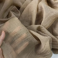A close-up of a person holding Longan Craft's Wrinkled Lyocell Fabric by the yard, a light brown, sustainable and breathable material with visible threads and soft folds, ideal for tops.