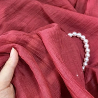A hand gently holds Longan Craft's wrinkled lyocell fabric by the yard in maroon, with a strand of white pearls resting on the breathable, solid color sustainable material perfect for tops.