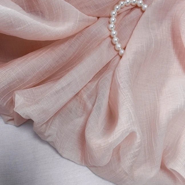 A strand of white pearls rests atop soft, pale pink Wrinkled Lyocell Fabric By The Yard from Longan Craft—a sheer, breathable, sustainable textile with gentle folds.