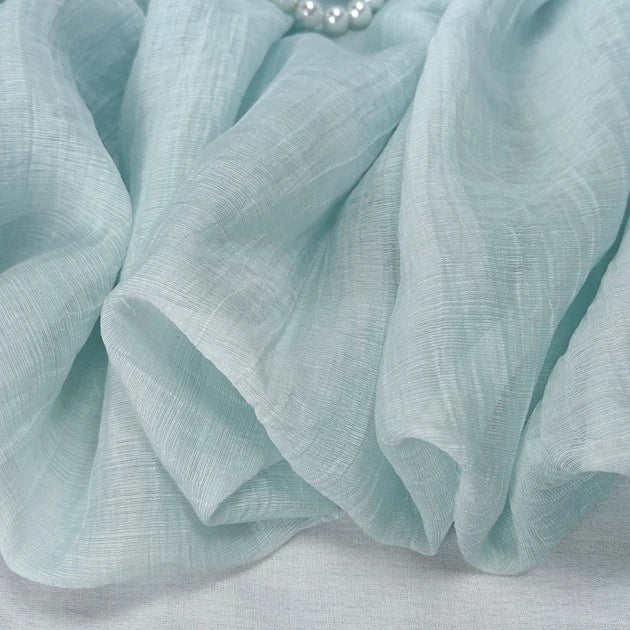 Close-up of Longan Craft's wrinkled light blue lyocell fabric, a sheer and breathable sustainable textile with gentle folds. A small strand of white pearls appears at the top. Ideal for tops, sold by the yard.