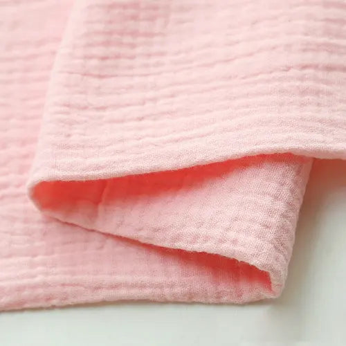 A close-up of soft pink Solid Color Cotton Double Gauze Fabric by the yard from Longan craft, featuring textured bubble gauze muslin ideal for quilting or making baby clothing, shown folded on a white surface.