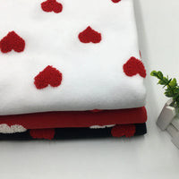 A folded stack of Longon Craft Heart Polar Fleece Fabric By The Yard, featuring 3D heart patterns on white, black, and red backgrounds, lies on a white surface beside a small green plant and two white books.