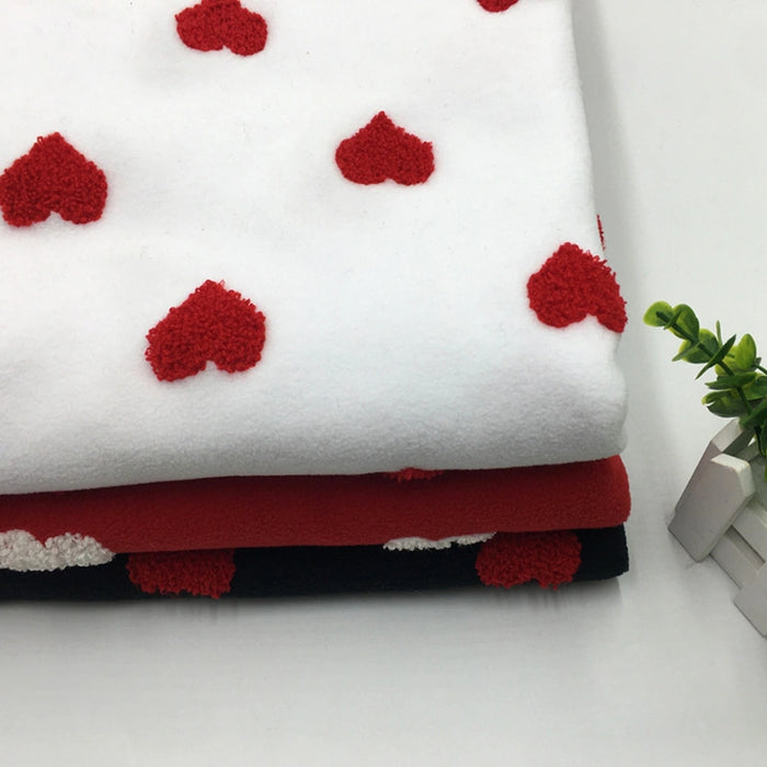 A folded stack of Longon Craft Heart Polar Fleece Fabric By The Yard, featuring 3D heart patterns on white, black, and red backgrounds, lies on a white surface beside a small green plant and two white books.