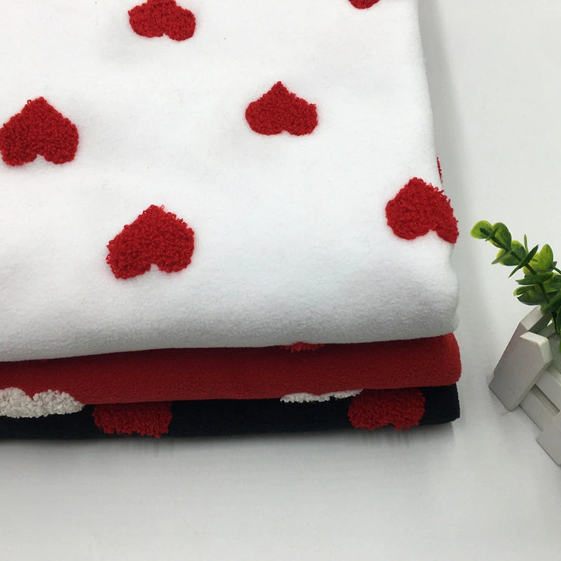 A folded stack of Longon Craft Heart Polar Fleece Fabric By The Yard, featuring 3D heart patterns on white, black, and red backgrounds, lies on a white surface beside a small green plant and two white books.