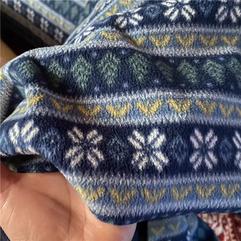 A hand holds Longon Craft’s Snowflake Polar Fleece Fabric By The Yard, a soft blue polyester with white snowflakes, yellow zigzags, and green diamond shapes—ideal for cozy DIY sewing projects.