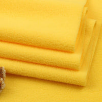 Four neatly folded yellow Longon Craft Solid Polar Fleece Fabric pieces—made of thick, plain 100% polyester—are stacked atop each other, highlighting their soft texture. A corner of another cozy fabric is visible in the bottom left.