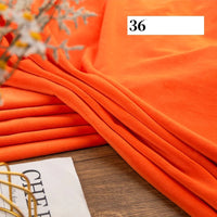 A piece of Longon Craft Stretch Minky Fabric By The Yard in bright orange is neatly folded on a wooden surface, with "36" shown in a white box. A magazine and plant are nearby—ideal for DIY clothing, plushies, dolls, pillows, and blankets.