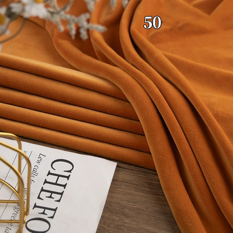 A stack of folded Stretch Minky Fabric by Longon Craft in an orange-brown shade rests on a wooden surface near a magazine and decor; the number 50 is visible on the fabric. Perfect for DIY clothing, pillow plushies, and dolls.