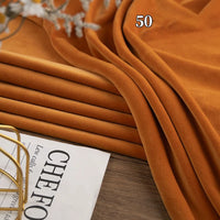 A stack of folded Stretch Minky Fabric by Longon Craft in an orange-brown shade rests on a wooden surface near a magazine and decor; the number 50 is visible on the fabric. Perfect for DIY clothing, pillow plushies, and dolls.