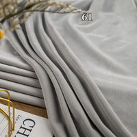A close-up of Longon Craft Stretch Minky Fabric By The Yard in light grey (#61), draped in folds, with a book and gold-rimmed glasses visible in the corner. Ideal for DIY clothing, plushies, pillows, and dolls.
