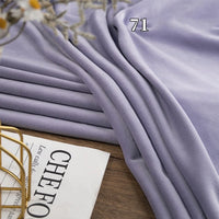 A folded piece of Longon Craft Stretch Minky Fabric By The Yard in light purple (#71) is displayed beside a magazine and decorative gold wire basket on a wooden surface. Ideal for DIY clothing, pillow, plushies, and dolls.
