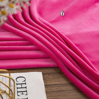 A stack of vibrant pink Longon Craft Stretch Minky Fabric lies on a wooden surface, perfect for DIY clothing, pillows, plushies, or dolls. Part of a magazine and some flowers are visible at the edge.
