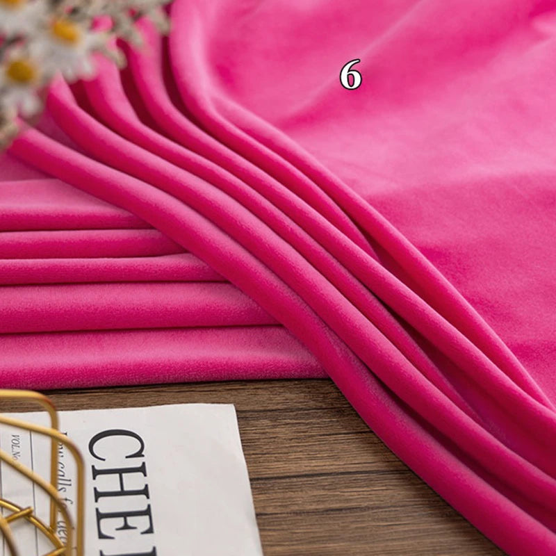 A stack of vibrant pink Longon Craft Stretch Minky Fabric lies on a wooden surface, perfect for DIY clothing, pillows, plushies, or dolls. Part of a magazine and some flowers are visible at the edge.