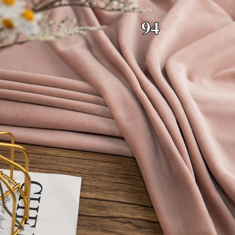 A folded piece of light pink Longon Craft Stretch Minky Fabric By The Yard is draped on a wooden surface beside a magazine and a decorative metal item.