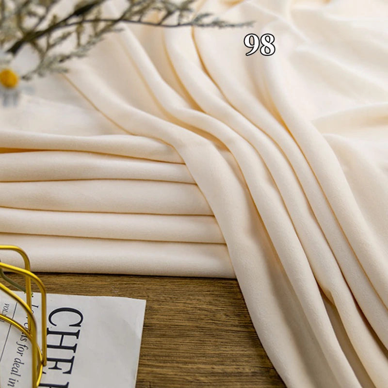 A folded piece of Longon Craft Stretch Minky Fabric in light beige is displayed on a wooden surface with the number 98; part of a magazine and eyeglasses can be seen in the corner.