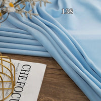 A folded piece of Longon Craft Stretch Minky Fabric By The Yard in light blue rests on a wooden surface near a magazine and decor item; the number 138 appears above the multicolor four-way stretch cuddle minky material.