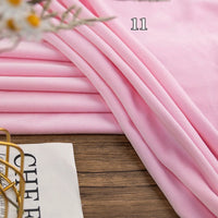 Light pink Stretch Minky Fabric by the yard from Longon Craft, a multicolor four-way stretch cuddle material for DIY clothing, pillows, plushies, and dolls, is neatly folded on a wooden surface with a partial magazine and flowers nearby.