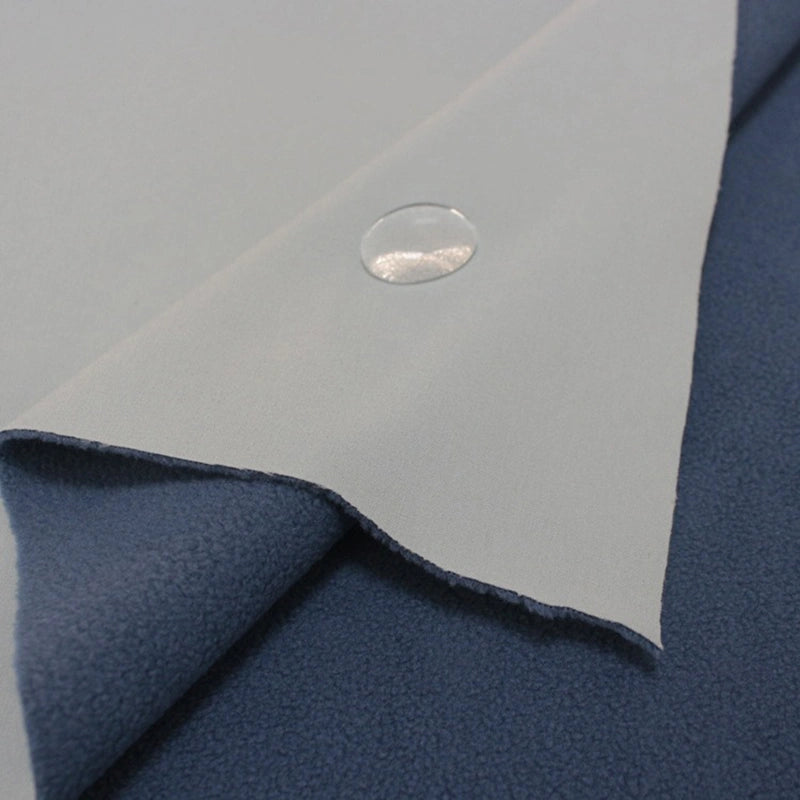 A close-up of Longon Craft Waterproof Polar Fleece Fabric By The Yard shows its water-resistant, light gray surface with a water droplet, layered over dark blue fleece. This 4 way stretch polyester fabric is ideal for sewing and crafts.
